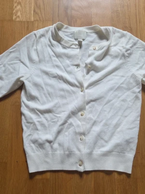 Vit kortärmad cardigan - Snygg vit cardigan med korta ärmar och rund halsringning. Tröjan har knappar framtill och är i ett tunt, mjukt material som känns skönt mot huden. Perfekt att bära öppen eller stängd för en clean och enkel look.