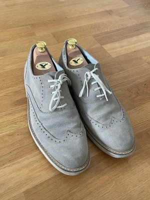 Grå mocka loafers med broguemönster - Snygga loafers i ljusgrå mocka med klassiskt broguemönster och vita snören. Skorna har en robust sula med kontrastsöm och elegant vingeformad tå. Perfekta för dig som vill ha en stilren och trendig look.
