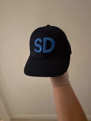 Svart keps med blå SD-brodyr - Snygg svart keps med böjd skärm och broderade bokstäver 'SD' i blått framtill. Justerbar rem baktill för perfekt passform. Kepsen är tillverkad i ett klassiskt bomullsmaterial och har en stilren look.