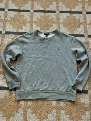 Grå sweatshirt från Polo Ralph Lauren - Klassisk grå sweatshirt från Polo Ralph Lauren med rund halsringning och diskret broderad logga på bröstet. Tröjan har ribbade muddar vid ärmslut och nederkant, perfekt för en avslappnad och stilren look.