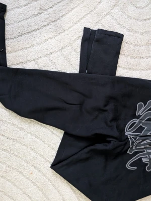 Black Sweatpants with Syna print - Svarta mjukisbyxor från LRG med stor grafisk print på ena benet. Tillverkade i mjukt, fleecefodrat material som känns skönt mot huden. Perfekta för en avslappnad streetwear-look.