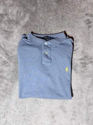 Blå pikétröja från Ralph Lauren - Snygg blå pikétröja från Ralph Lauren med klassisk krage och två knappar framtill. Tröjan har det ikoniska gula broderade logotypen på bröstet och är i bomull. Perfekt för en stilren och avslappnad look.