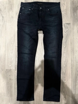 Nudie Jeans Tape Ted Mörkblå/Svarta W33/L32 - Slim Fit - Nudie Jeans Tape Ted ”Blue Black” mörkblå/svarta jeans med slim fit-passform, normalhög midja och avsmalnande ben, storlek W33/L32, midja 42 cm, ytterbenslängd 108 cm, benöppning 18,5 cm, modellen är 185cm & väger 65kg, i fint skick.