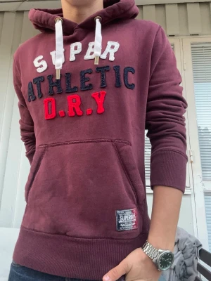 Vintage Superdry Hoodie med Nummer - En av de fetaste hoodies jag någonsin sett, av just det trendiga märket Superdry🤩 Istället för tryck har man på denna hoodie valt att jobba med brodyr och tyg för att kvalitén och utseendet ska märkas🙌 På baksidan har vi en numret ”23” i brodyr, vilket verkligen färdigställer denna extremt snygga hoodie.   Om den inte passat mig ytterst lite långt, hade denna hoodie vart längst fram i garderoben. Stl M, Athletics serien.🤩  Färg: Vinröd🍷 Längd från axel: 67cm.          Bredd mellan armhålor: 52cm