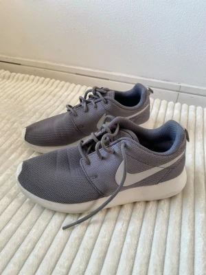 Grå Nike sneakers med vit sula - Snygga grå sneakers från Nike med vit sula och klassisk swoosh-logga på sidan. Skorna har ovandel i meshmaterial för bra ventilation och snörning framtill. Perfekta för en sportig och avslappnad stil.