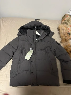 Mörkgrå dunjacka från Canada Goose - Snygg mörkgrå dunjacka från Canada Goose med avtagbar huva och klassisk logotyp på ärmen. Jackan har dragkedja och knappar framtill, två stora fickor och varm vaddering. Perfekt för kalla vinterdagar.