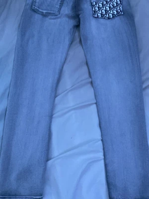 Ljusblå raka jeans med unik ficka - Säljer ett par ljusblå raka jeans med en cool detalj – ena bakfickan har ett vitt tyg med svart mönster. Jeansen har klassisk passform och är tillverkade i denim. Perfekta för dig som vill sticka ut lite extra!
