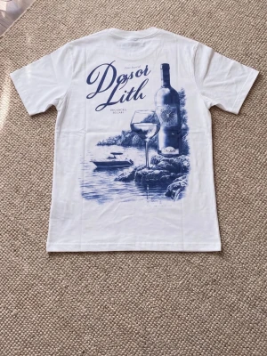 Vit t-shirt med blått tryck - Säljer en vit t-shirt med ett stort blått tryck på ryggen som föreställer en vinflaska, ett vinglas och en havsmiljö. T-shirten har korta ärmar och klassisk passform. Perfekt för dig som gillar stilrena motiv och vill sticka ut lite extra.