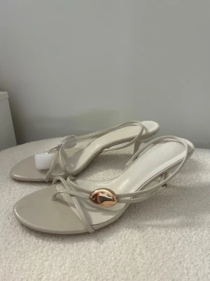 Beiga klackar med gulddetalj - Snygga sandaletter i beige med tunna remmar och en elegant guldfärgad detalj framtill. Sandalerna har öppen tå och låg klack, vilket ger en stilren och modern look. Tillverkade i ett glansigt material som ger en lyxig känsla. Helt oanvända!💕