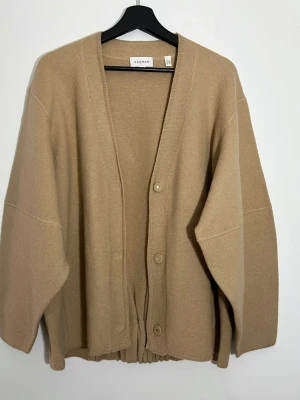 Beige oversized kofta från Dagmar - stl S - Beige oversized kofta från Dagmar med v-ringning och knäppning framtill. Koftan har breda ärmar och ribbad detalj i midjan baktill. Tillverkad i mjukt stickat material som ger en avslappnad och trendig look.