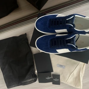 Blå och vita sneakers från Valentino - Snygga sneakers i blå mocka och vitt läder från Valentino. Skorna har klassisk låg profil, vita detaljer på sidorna och matchande vita sulor. Perfekta för dig som vill ha en stilren och trendig look.