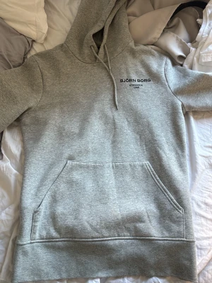 Grå hoodie från Björn Borg - Snygg grå hoodie från Björn Borg med klassisk känguruficka och justerbar huva. Diskret logga på bröstet och mjukt material som känns skönt mot huden. Perfekt för en avslappnad och sportig stil.