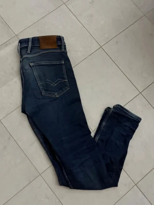 Mörkblå slim fit jeans från Replay - Snygga mörkblå slim fit jeans från Replay med klassiska fem fickor och detaljerade sömmar på bakfickorna. Jeansen har en läderpatch vid linningen och subtila slitningar för en trendig look. Perfekta för dig som gillar stilrena och moderna jeans.