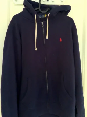 Mörkblå hoodie med dragkedja - Mörkblå hoodie med dragkedja från Polo Ralph Lauren. Tröjan har vit dragsko i huvan, två fickor framtill och en röd broderad logga på bröstet. Perfekt för en avslappnad och stilren look.