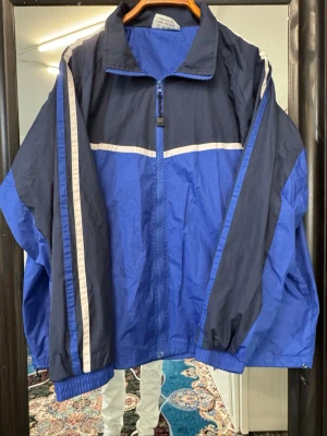 Blå och marinblå retro vindjacka - Snygg retro vindjacka i blått och marinblått med vita detaljer. Jackan har dragkedja framtill, ståkrage och elastiska muddar vid ärmarna. Lätt material som passar perfekt till streetwear och sportig stil. Två sidofickor med dragkedja.