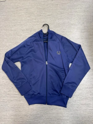 Blå ziptröja från Fred Perry - Snygg blå ziptröja från Fred Perry med ribbade muddar och krage. Tröjan har dragkedja framtill, två fickor och den klassiska Fred Perry-loggan broderad på bröstet. Tillverkad i ett glansigt, sportigt material som ger en retrovibe.