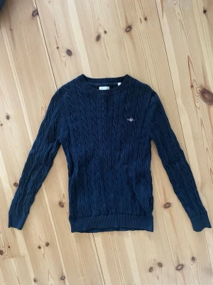 Mörkblå kabelstickad tröja från Gant - Mörkblå kabelstickad tröja med rund halsringning och ribbade muddar vid ärmslut och nederkant. Tröjan har ett diskret broderat emblem på bröstet och är långärmad. Perfekt för lager på lager och en klassisk look.