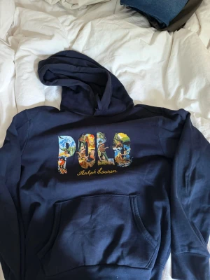 Mörkblå hoodie från Polo Ralph Lauren - Snygg mörkblå hoodie från Polo Ralph Lauren med stor färgglad POLO-logga och broderad text på bröstet. Klassisk huva med dragsko och magficka framtill. Perfekt för en avslappnad och trendig stil.