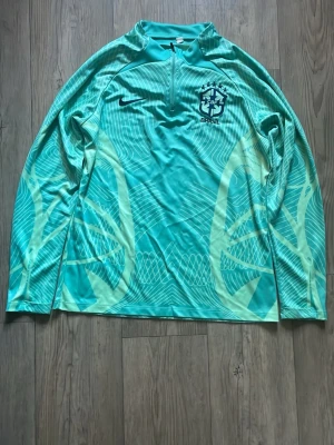Turkos Brasil långärmad träningströja - Turkos långärmad träningströja från Nike med Brasil-logga på bröstet. Tröjan har halv dragkedja vid halsen och ett grafiskt mönster i ljusgult över hela plagget. Tillverkad i ett lätt och snabbtorkande material som passar perfekt till träning.