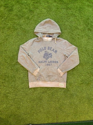 Grå hoodie Polo Bear Ralph Lauren storlek s passar perfekt om man är 178cm - Säljer en grå hoodie från Ralph Lauren med Polo Bear-tryck och texten 'Ralph Lauren 1967' framtill. Hoodien har huva med dragsko, ribbade muddar och en patch nertill. Mjuk insida och klassisk passform, perfekt för en avslappnad stil.