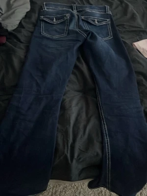 Mörkblå bootcut jeans med kontrastsömmar - Säljer ett par mörkblå bootcut jeans med vita kontrastsömmar och fem fickor. Jeansen har breda ben och klassisk knappgylf. Perfekta för dig som gillar en retro vibe och vill ha ett par jeans som sticker ut lite extra.
