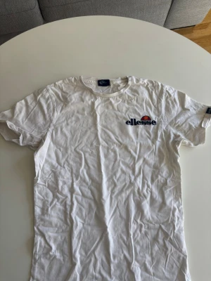 Vit t-shirt från Ellesse - Säljer en klassisk vit t-shirt från Ellesse med logga i blått och orange på bröstet. T-shirten har rund hals och korta ärmar, tillverkad i mjuk bomull för en skön känsla. Perfekt till jeans eller shorts.