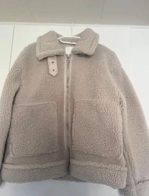 Beige teddyjacka med krage - Mysig beige teddyjacka med stor krage och dekorativt spänne. Jackan har två stora framfickor, dragkedja framtill och breda muddar vid ärmslut och nederkant. Perfekt för kalla dagar och ger en trendig look.