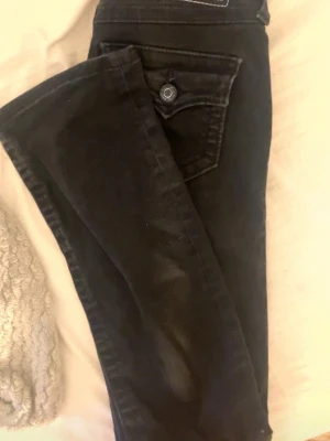 Svarta bootcut jeans True religion - Säljer ett par svarta bootcut jeans, True Religion, med snygga detaljer på fickorna och klassisk knappstängning. Jeansen har en skön passform och är tillverkade i ett mjukt denim-material som sitter bra på kroppen. Finns små små defekter men inte märkbart på långt håll.