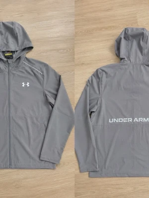 Grå vindjacka från Under Armour - Snygg grå vindjacka från Under Armour med huva och dragkedja framtill. Jackan har logga på bröstet och stor text på ryggen. Tillverkad i lätt och vindtätt material, perfekt för blåsiga dagar. Enkel och stilren design med diskreta detaljer.