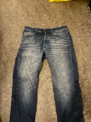 Jeans från Jack and Jones 29/30 - Säljer ett par mörkblå jeans med snygga slitningar och tvättade detaljer på låren och knäna. Klassisk femficksmodell med knapp och dragkedja. Jeansen har raka ben och är tillverkade i denim med en cool, avslappnad look.