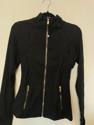 Lululemon 4,6,8  - Snygg svart jacka med figurnära passform och guldfärgade dragkedjor framtill och på fickorna. Jackan har hög krage och långa ärmar, perfekt för en sportig och stilren look. Materialet känns mjukt och stretchigt.