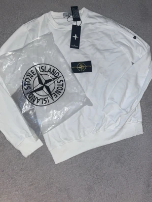 Vit sweatshirt från Stone Island - Säljer en stilren vit sweatshirt från Stone Island med klassisk rund hals och ribbade muddar. Tröjan har diskret logotyp på ärmen och levereras med originalpåse och taggar. Perfekt för dig som gillar streetwear och enkel design.