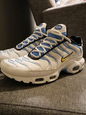 Nike Air max  - Nike Air Max sneakers i vitt och blått med svarta och gula detaljer. Skorna har en mesh- och syntetöverdel, synlig Air-dämpning i sulan och den ikoniska TN-loggan på hälen. Snyggt vågmönster och klassisk siluett.