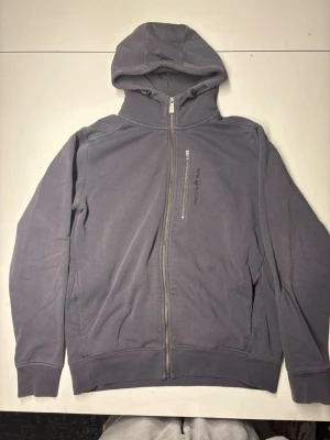 Mörkblå hoodie med dragkedja - Säljer en lila hoodie med hel dragkedja framtill och huva. Tröjan har en bröstficka med dragkedja och diskret tryck på bröstet. Perfekt för en avslappnad och cool stil. Materialet känns mjukt och bekvämt mot huden.