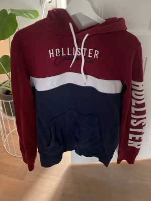 Hollister hoodie röd och marinblå M - Snygg hoodie från Hollister i rött och marinblått med vit detalj. Stor logga på bröstet och ärmen, samt känguruficka och huva med vita snören. Perfekt för en avslappnad och trendig look.