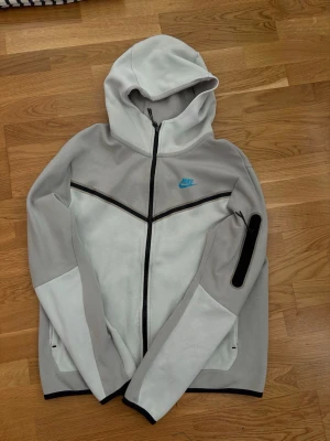 Nike grå hoodie med dragkedja - Nike hoodie i ljusgrått och vitt med svart dragkedja och svarta detaljer. Har huva, blå Nike-logga på bröstet och en praktisk ficka med dragkedja på ärmen. Tillverkad i mjukt material, perfekt för en sportig och avslappnad stil.