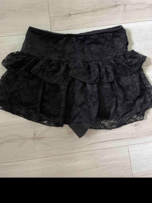 Svart spetsig skort med volanger - Supersnygg svart skort med volanger och spetsdetaljer. Kjoldelen har ett lager av genombruten spets och volangkant som ger en feminin och trendig look. Perfekt för dig som vill ha kjolkänsla men ändå shorts under. Materialet är lätt och stretchigt. Lite oklart vart den köptes, men den användes första natten på the weeknd i Stockholm