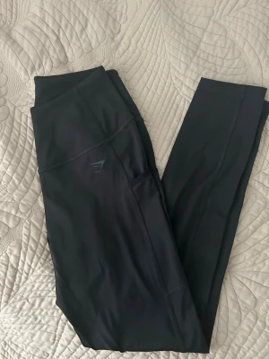 Svarta leggings från Gymshark - Snygga svarta leggings från Gymshark med hög midja och diskret logga på benet. Tillverkade i ett stretchigt och mjukt material som passar perfekt till träning eller chill. Har praktisk ficka på sidan för mobilen. Superskön passform och stilren look.
