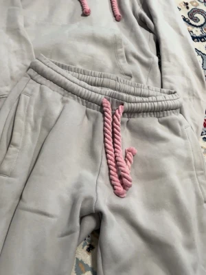 Grå mjukisset med rosa detaljer och graffiti-tryck - Säljer ett grått mjukisset med rosa snören och ett stort rosa graffiti-tryck på baksidan av byxorna. Setet består av en hoodie och ett par mjukisbyxor. Perfekt för vardag eller mys hemma! Kontakta för mer info eller prisförslag.