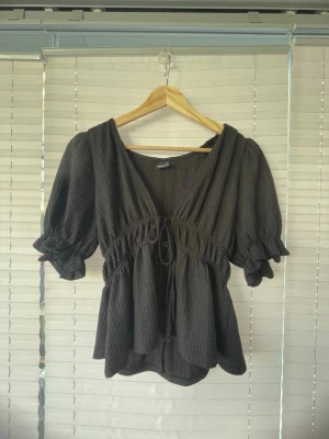Svart blus med puffärm och knyt - Säljer en svart blus med puffärmar och v-ringning. Toppen har knyt i midjan och volangdetaljer nedtill. Materialet är mjukt och har en lätt struktur som ger en avslappnad känsla. Perfekt för dig som gillar en feminin och trendig look.
