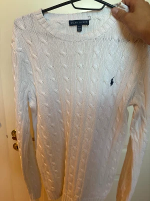 Vit kabelstickad tröja Ralph Lauren - Kabelstickad vit tröja från Ralph Lauren med rund halsringning och lång ärm. Klassisk marinblå logga broderad på bröstet. Tröjan är i mjukt material och har ribbade muddar vid ärmslut och nederkant. Perfekt för en clean och stilren look.