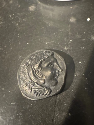 Antik grekisk mynt med motiv - Säljer ett antikt grekiskt mynt i metall med motiv av ett manshuvud på ena sidan och en sittande figur med stav och fågel på andra sidan. Myntet har text på grekiska och en mörk, något ojämn form. Perfekt för dig som gillar historia eller samlar på gamla mynt.