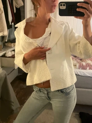 Vit jacka med korta ärmar - Stilren vit kavaj med bred krage och två knappar framtill. Kavajen har unika spetsdetaljer vid ärmslut och nederkant som ger en romantisk touch. Passar perfekt till jeans eller kjol för en trendig look. Materialet ser ut att vara en blandning av bomull och spets. Lite små fläckar på kragen men inget man tänker på. Vet ej storlek men passar xs-m