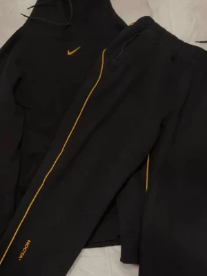 Svart Nike NOCTA tracksuit set - Säljer ett svart tracksuit set från Nike NOCTA med gul logga och detaljer. Setet består av en hoodie med dragsko och matchande byxor med gul linje längs benet och NOCTA-tryck vid ankeln. 