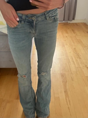Ljusblå bootcut jeans med slitningar - Säljer ett par ljusblå bootcut jeans med slitningar vid knäna och snygga broderade detaljer på bakfickorna. Jeansen har låg midja och klassisk femficksdesign. Perfekta för en avslappnad och trendig look.