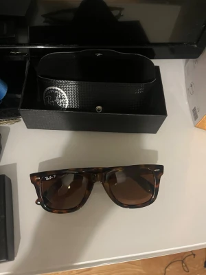 Ray-Ban bruna wayfarer solglasögon - Säljer ett par klassiska Ray-Ban wayfarer solglasögon i brunmelerad färg. De har en fyrkantig form och kommer med originalfodral och box. Perfekta för dig som vill ha en tidlös och snygg accessoar till sommaren.