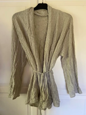 Beige crinklad strandpullover med knytband - Luftig och mjuk strandpullover i beige med crinklad struktur och långa ärmar. Modellen har ett smalt knytband i midjan och öppen front utan knappar.
