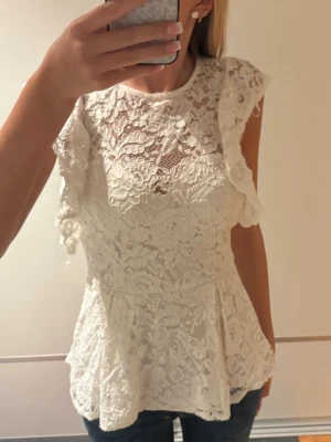 Vit spetsblus med volangdetaljer - Säljer en superfin vit blus i spets med volangdetaljer på ärmarna och rund hals. Blusen har en lätt utsvängd passform och knapp i nacken. Perfekt för dig som gillar romantisk stil och vill ha något extra till garderoben.