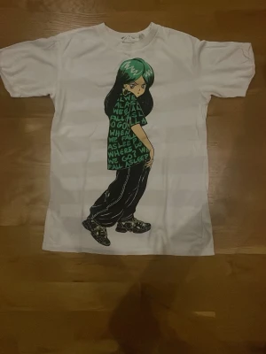 Vit t-shirt med anime-tryck från H&M - Vit t-shirt från H&M med ett stort anime-inspirerat tryck på framsidan. Motivet visar en karaktär med grönt hår och text på tröjan. T-shirten har rund hals och korta ärmar, perfekt för dig som gillar streetstyle och grafiska prints. VID ARMEN HAR SÖMMEN GÅTT UPP LITE MAN KAN SE PÅ BILD 3!! OXH DÄRFÖR HAR JAG LAGT UPP DEN BILLIGT!