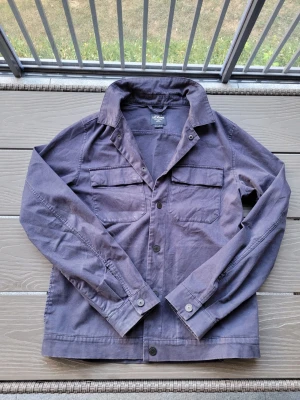 Lila overshirt med fickor - Stilren lila overshirt i bomull med två bröstfickor och knappar framtill. Klassisk krage och raka ärmar med knapp vid ärmslut. Perfekt lager-på-lager-plagg för en avslappnad look. Har tyvär fått en sminkfläck på ryggen som dock inte syns vid användning(endast om man kollar noga). Skulle tro att den går att få bort om det önskas